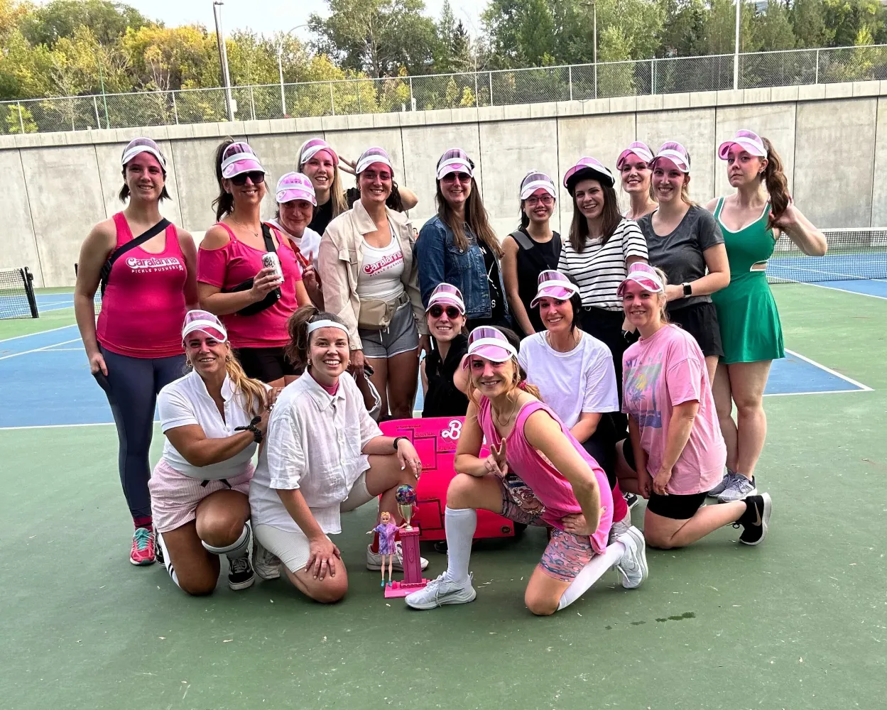 A group of women posing together on an outdoor tennis court, dressed in pink-themed athletic outfits and accessories. Many are wearing pink visors and have pink face paint. Some are kneeling in the front while others stand in the back, all smiling at the camera. A pink trophy and Barbie dolls are placed on the ground in front of them. The setting is an outdoor sports facility with a concrete wall and trees in the background.