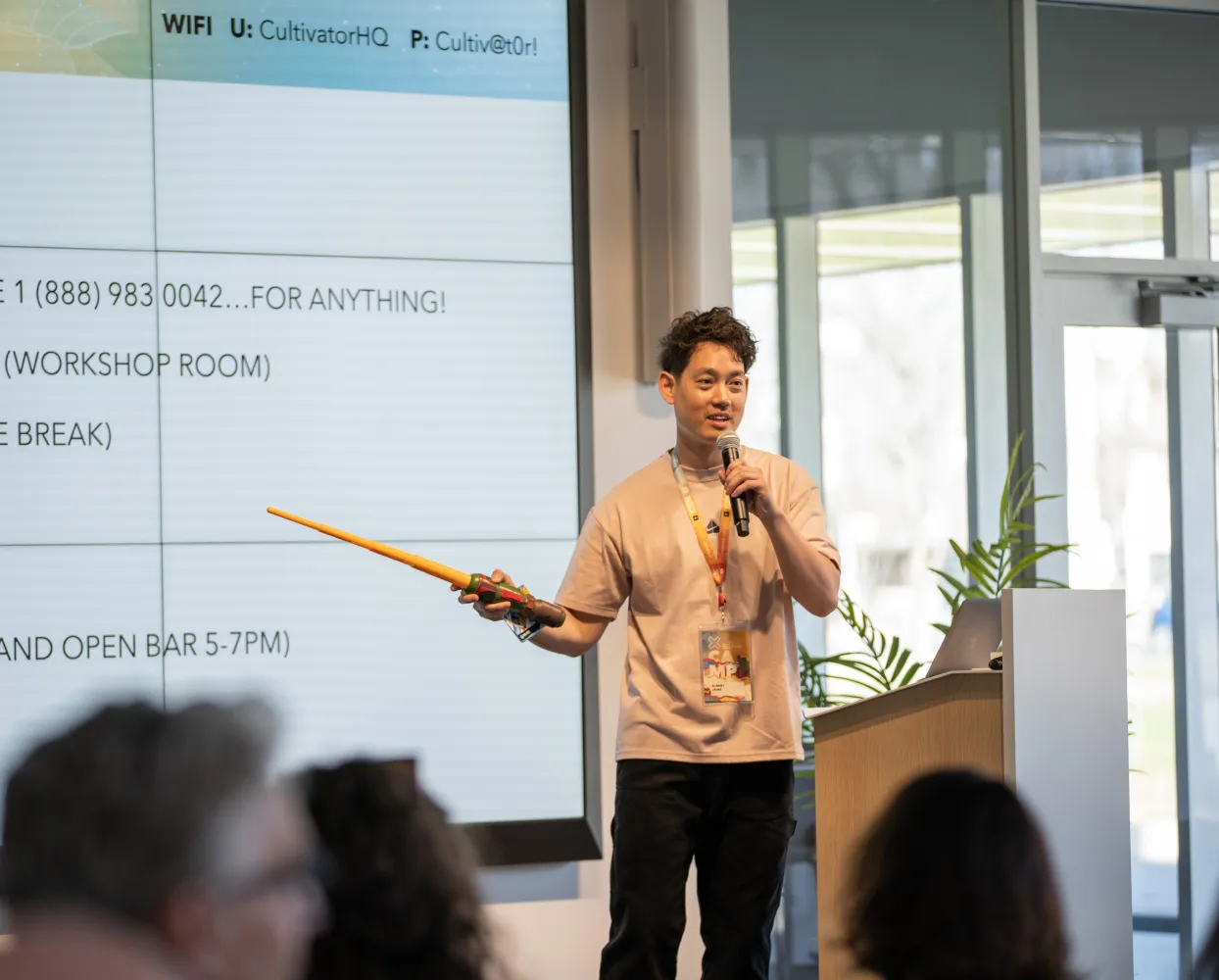 A young man with short curly hair is giving a presentation in a modern conference room. He is holding a microphone in one hand and a toy lightsaber in the other while speaking to an audience. He wears a beige t-shirt, a lanyard with a name badge, and dark pants. A large screen behind him displays text and a WiFi login. There is a wooden podium with a laptop on it, and large windows letting in natural light. The audience is partially visible in the foreground.