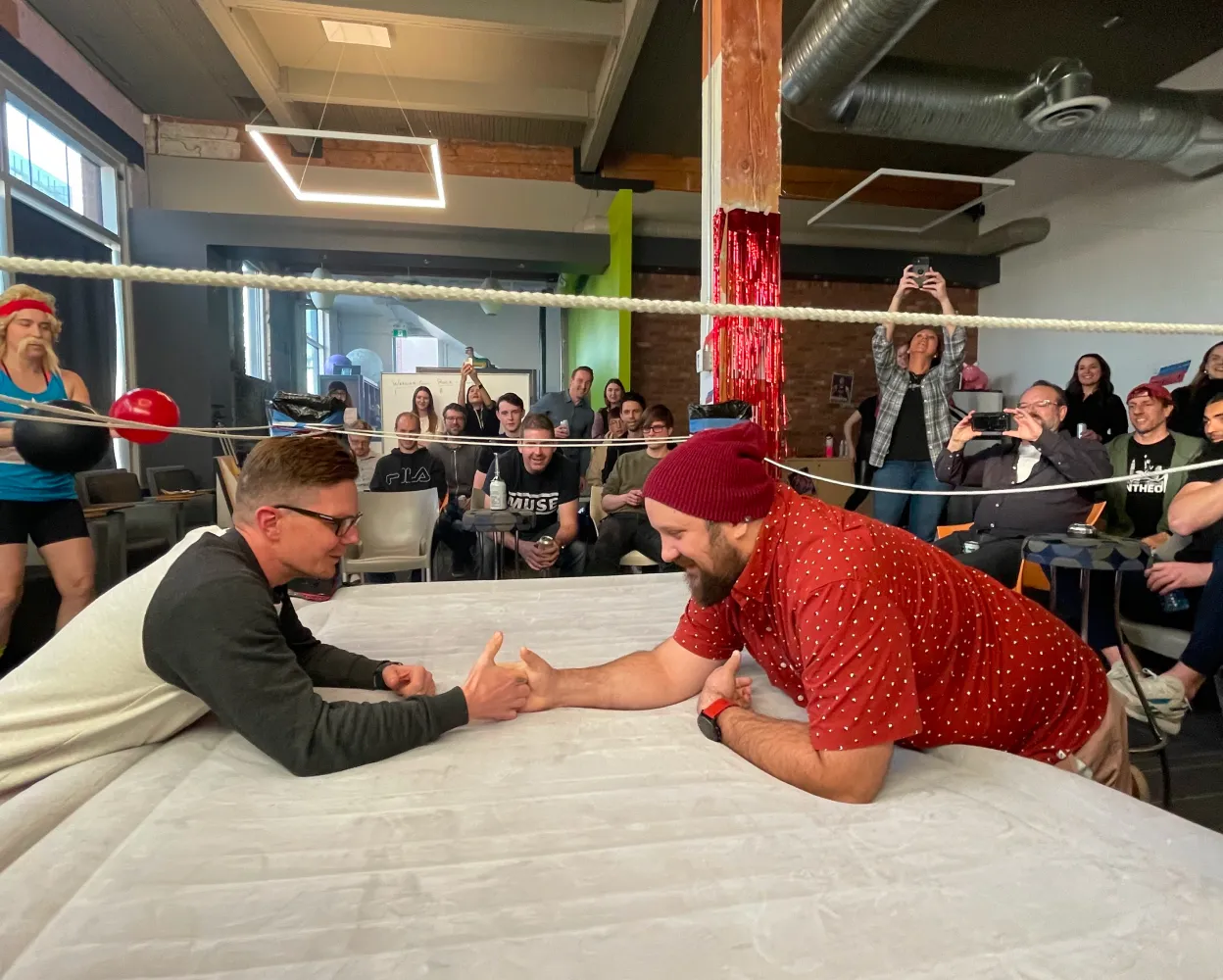 Two men are engaged in a thumb wrestling match inside a makeshift wrestling ring with a white mat and red rope decorations. One man wears glasses and a black-and-white long-sleeve shirt, while the other wears a red polka-dot shirt and a maroon beanie. They are smiling and laughing as they compete. A crowd of spectators surrounds them, cheering and taking photos. In the background, a person dressed as a boxer with red gloves and a headband stands near the ring. The setting is an indoor event space with industrial-style ceilings, exposed ductwork, and modern lighting.