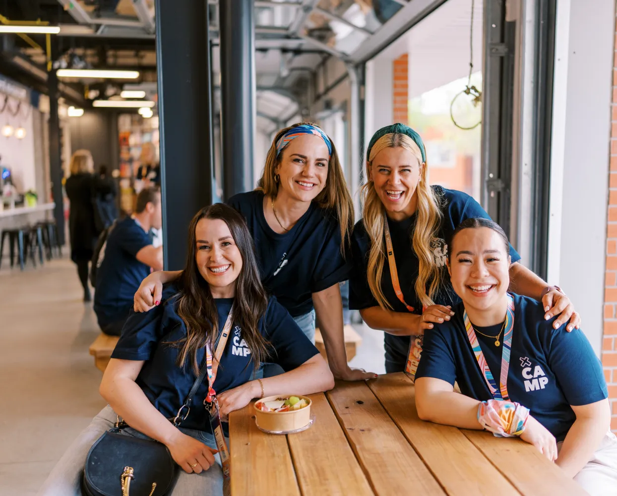 Four women wearing matching navy blue 'X-CAMP' t-shirts are gathered around a wooden picnic table, smiling at the camera. They are wearing colorful lanyards and some have headbands or scrunchies. One woman has a bowl of fruit in front of her. The setting is a modern, open-concept space with large windows, exposed metal beams, and a casual seating area. Other people are visible in the background, engaging in conversation.