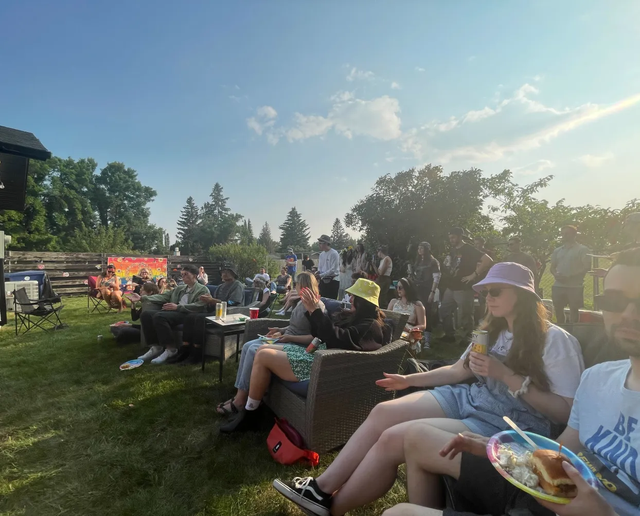 A large group of people is gathered outdoors in a grassy backyard during a sunny day. Some are seated on patio furniture, while others stand in the background. Many are wearing hats, sunglasses, and casual summer clothing. Some people are holding drinks and plates of food, including sandwiches and coleslaw. A colorful backdrop with a vibrant design is visible in the background. The setting includes trees, a wooden fence, and a clear blue sky.