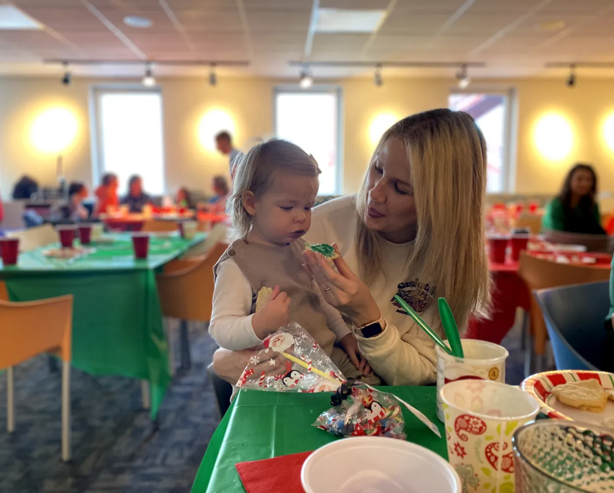 A young child with light-colored hair is sitting on a woman’s lap at a holiday-themed gathering. The woman, with long blonde hair, is holding a green frosted cookie and showing it to the child, who looks at it curiously while holding a piece of another cookie. The table in front of them has festive decorations, cups, and plates with Christmas-themed designs. The background features other people sitting at tables covered with red and green tablecloths in a warmly lit room.