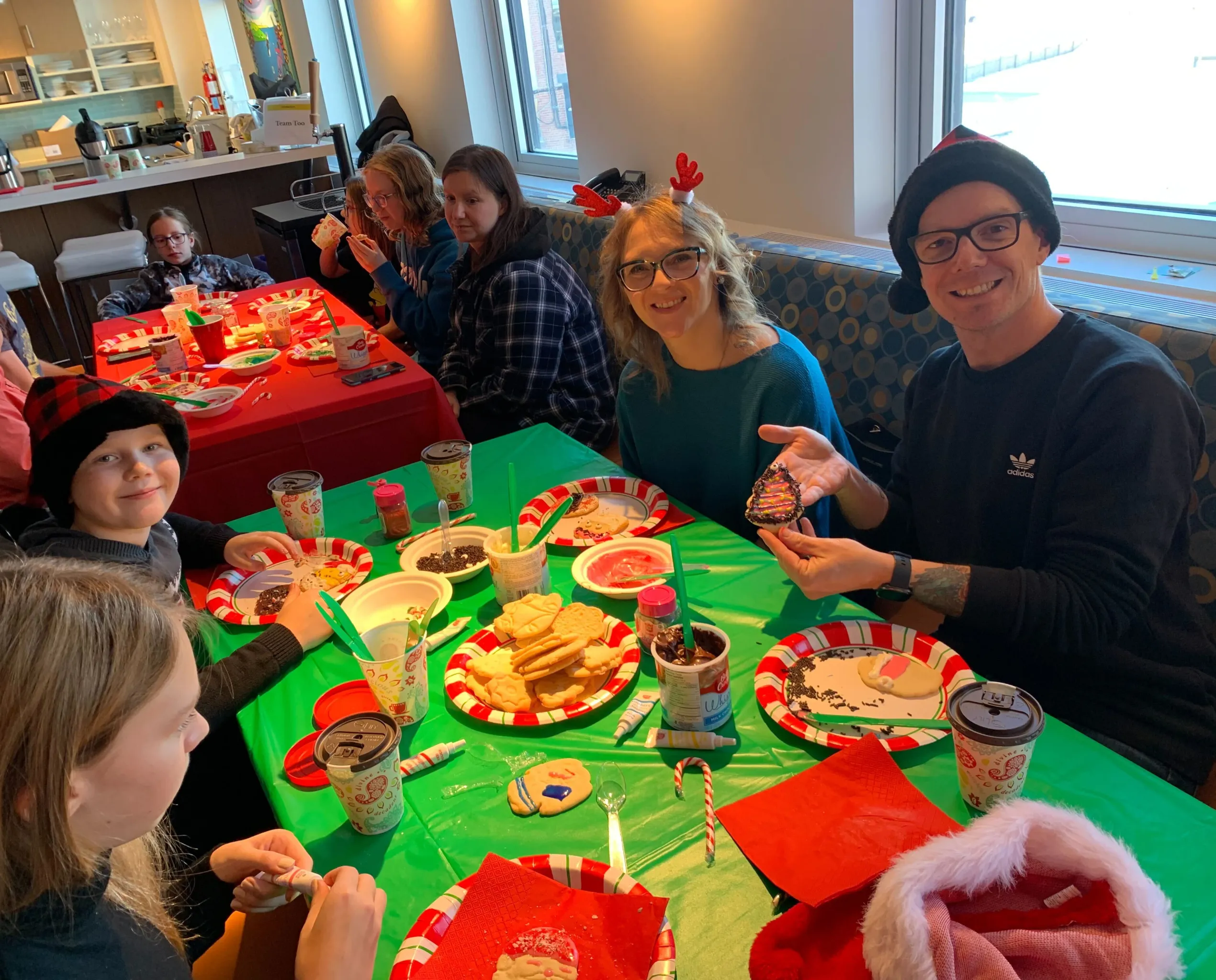 A group of people, including children and adults, is gathered around a table covered with a green tablecloth, decorating holiday cookies. The table is filled with Christmas-themed plates, icing, sprinkles, and cookies in various stages of decoration. A man wearing glasses and a black Adidas sweater is smiling and holding up a decorated Christmas tree-shaped cookie. A woman next to him, wearing a teal sweater and red reindeer antlers, is also smiling. A young boy in a red and black plaid hat grins at the camera. The setting is a festive indoor space with holiday decorations and a warm, cheerful atmosphere.