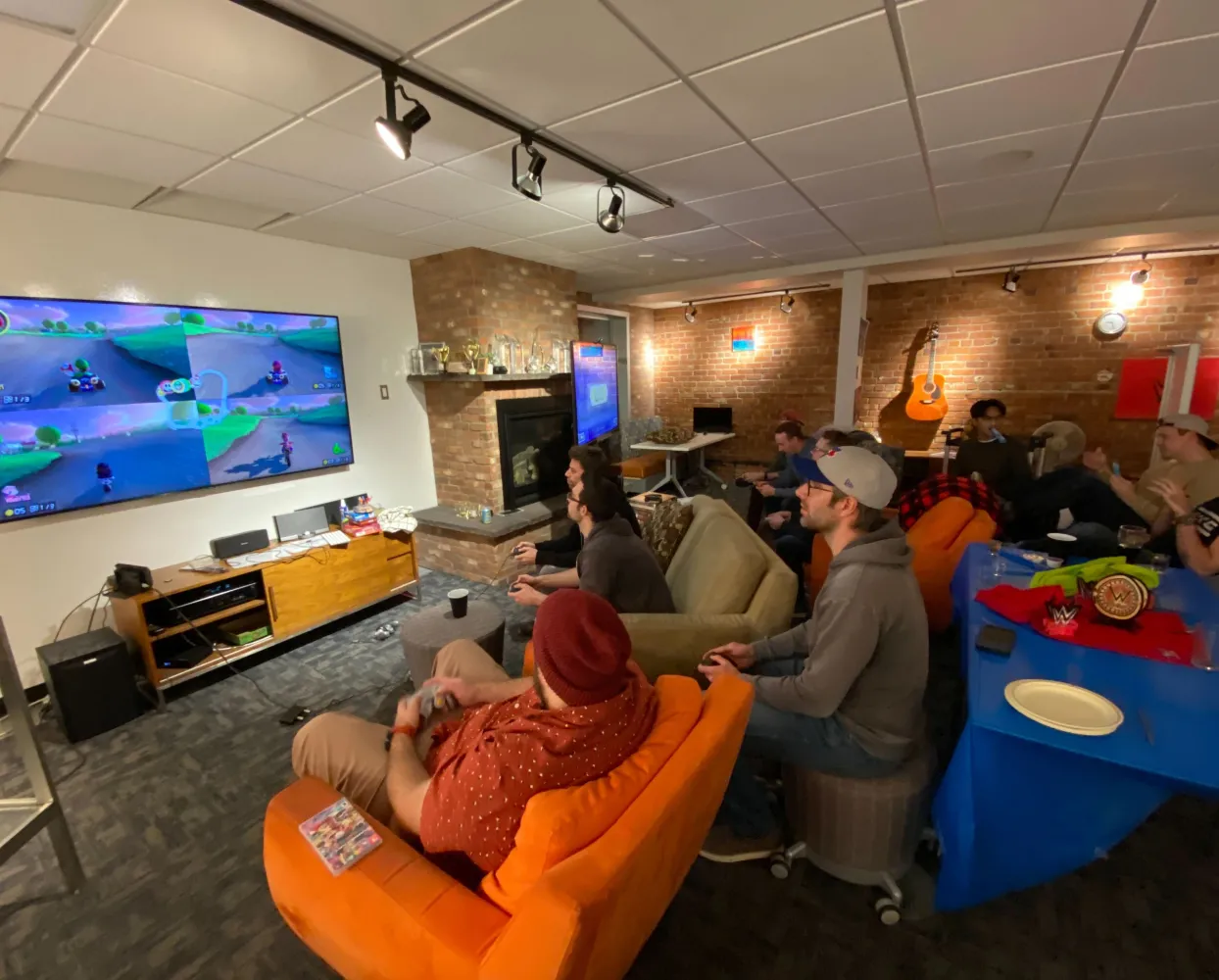 A group of people is gathered in a cozy lounge area playing a multiplayer racing video game on a large wall-mounted TV. Several players are seated on a mix of couches, bean bags, and stools, holding controllers and focusing on the game. The room has exposed brick walls, a fireplace, and a guitar hanging on the wall. A blue table nearby has wrestling championship belts and other items. Additional people are in the background, watching and socializing. The setting has a casual and fun atmosphere.