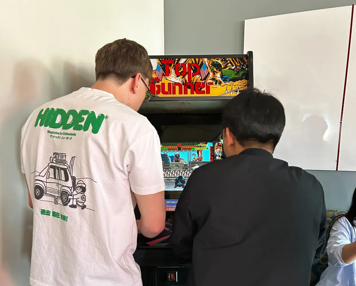 Two people are playing a retro arcade game on a 'Top Gunner' arcade machine. One person, wearing a white t-shirt with a green 'HIDDEN' logo and an illustration of a vehicle, is on the left, while the other, dressed in a black outfit, is on the right. They are focused on the game screen, which displays a classic side-scrolling action scene. The setting is indoors, with a white wall and a partially visible person sitting nearby.