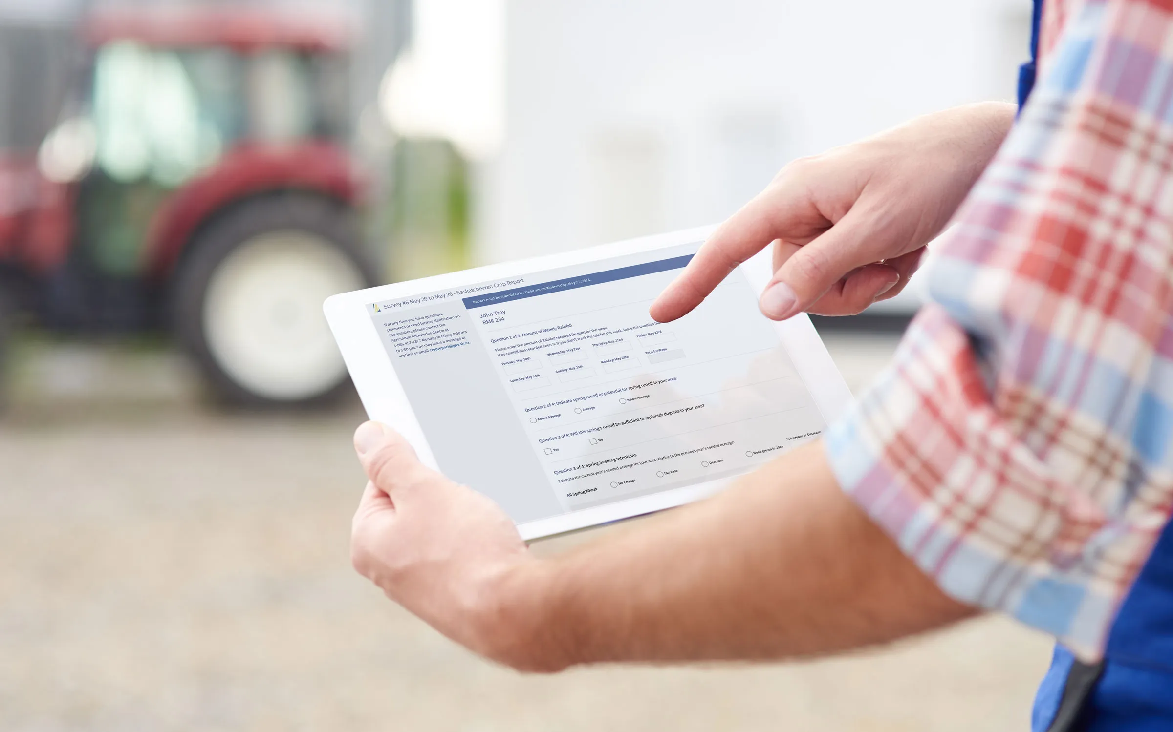 Farmer holding a tablet with a crop survey form, with a tractor blurred in the background