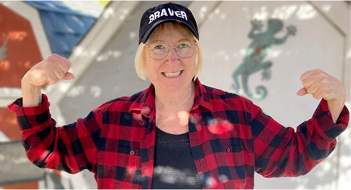 An image of Joanne Kachur wearing a hat that says 'braver', posing with her arms flexed and a smile on her face.