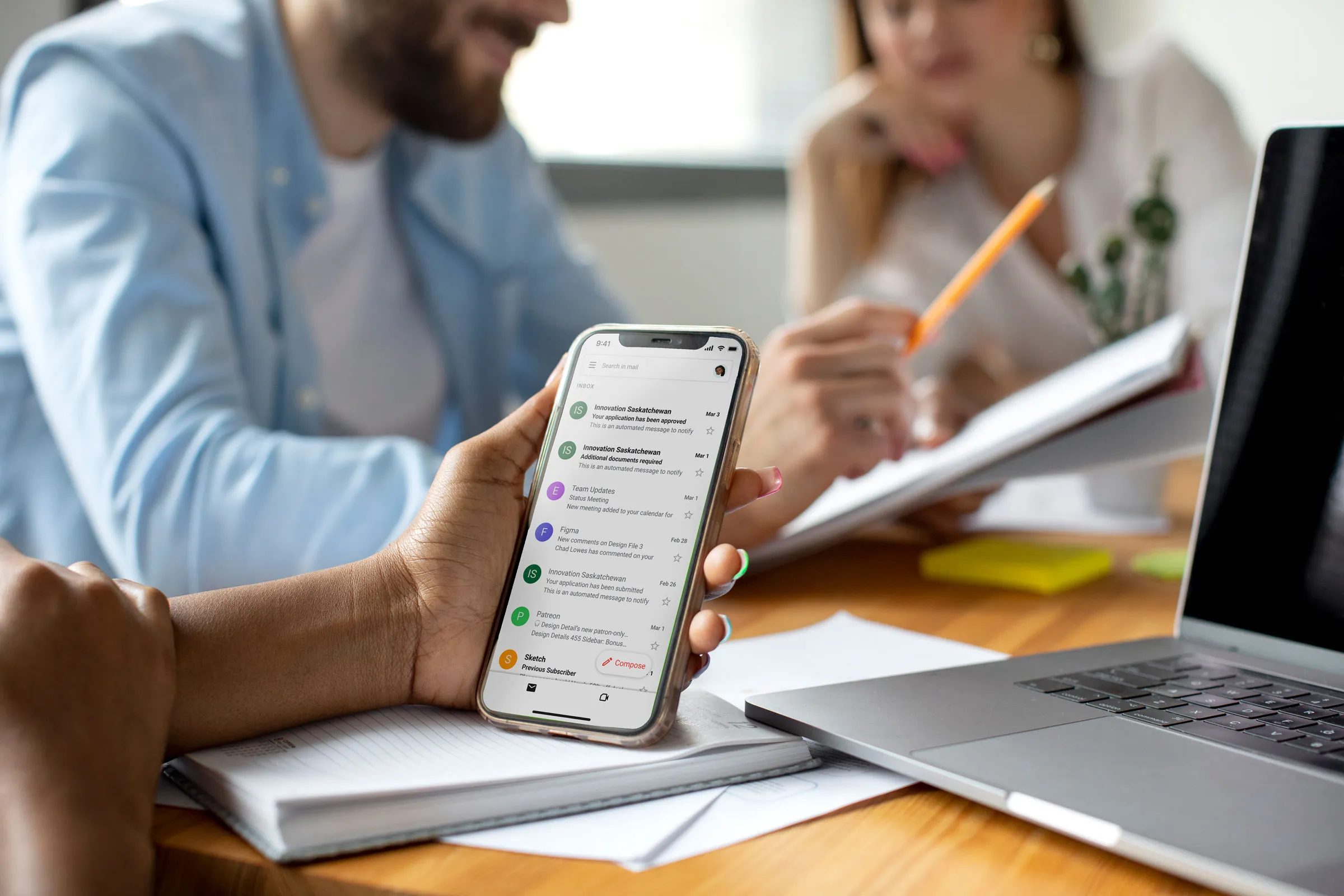 Person holding a smartphone showing Innovation Saskatchewan email updates during a meeting with a laptop nearby